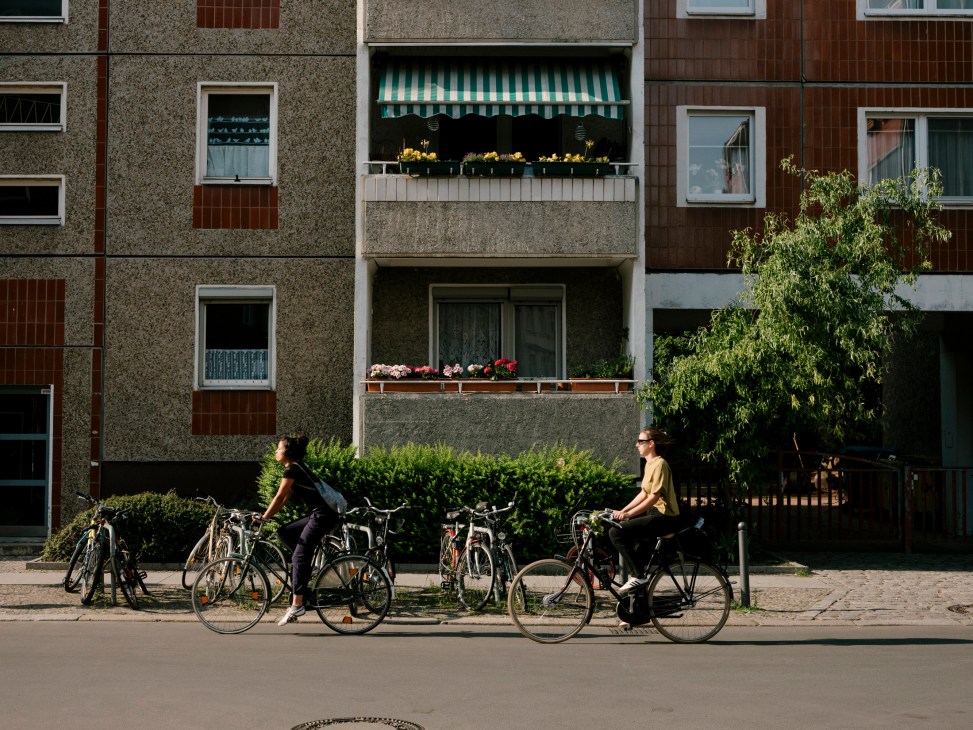 Two cyclists with bicycles parked in front of mid-century residential apartment buildings with flower-filled balconies.