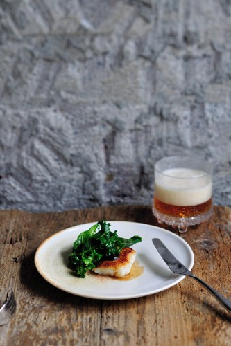 Plate with seared fish fillet and sautéed greens alongside a glass of beer on wooden table.