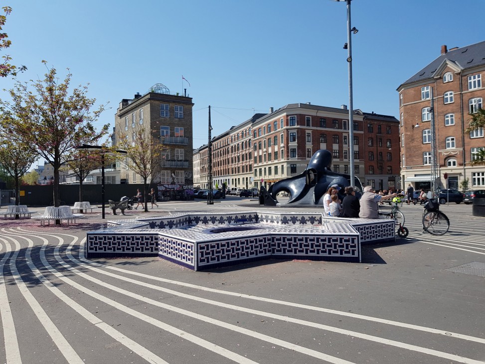 Public plaza with geometric patterned water feature, black sculptural art installation, and brick buildings in Copenhagen's Superkilen park.