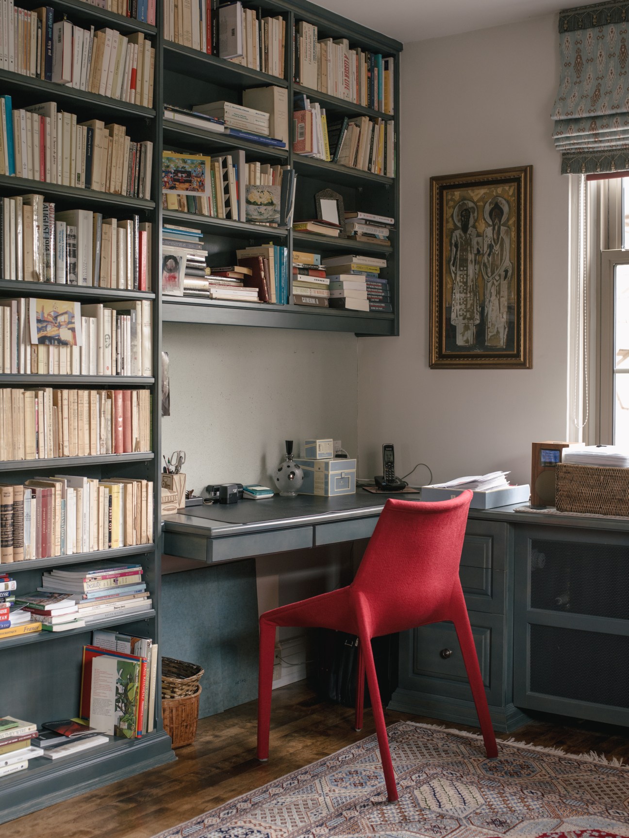 Home office with floor-to-ceiling dark shelving unit filled with books, red modern chair, and religious artwork on wall.