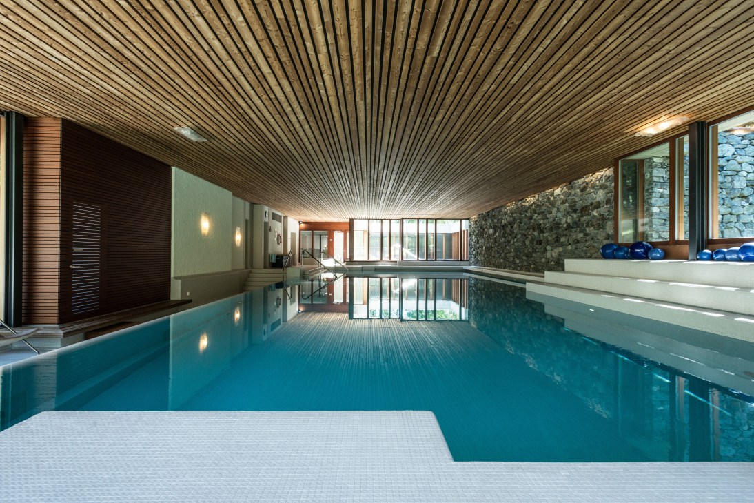 Indoor swimming pool with striped wooden ceiling, blue water, and glass-walled spa area at Hotel Waldhaus.
