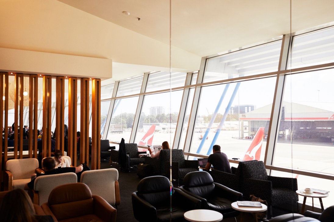 Qantas domestic business lounge interior with seating areas and large windows overlooking airport tarmac with aircraft visible.