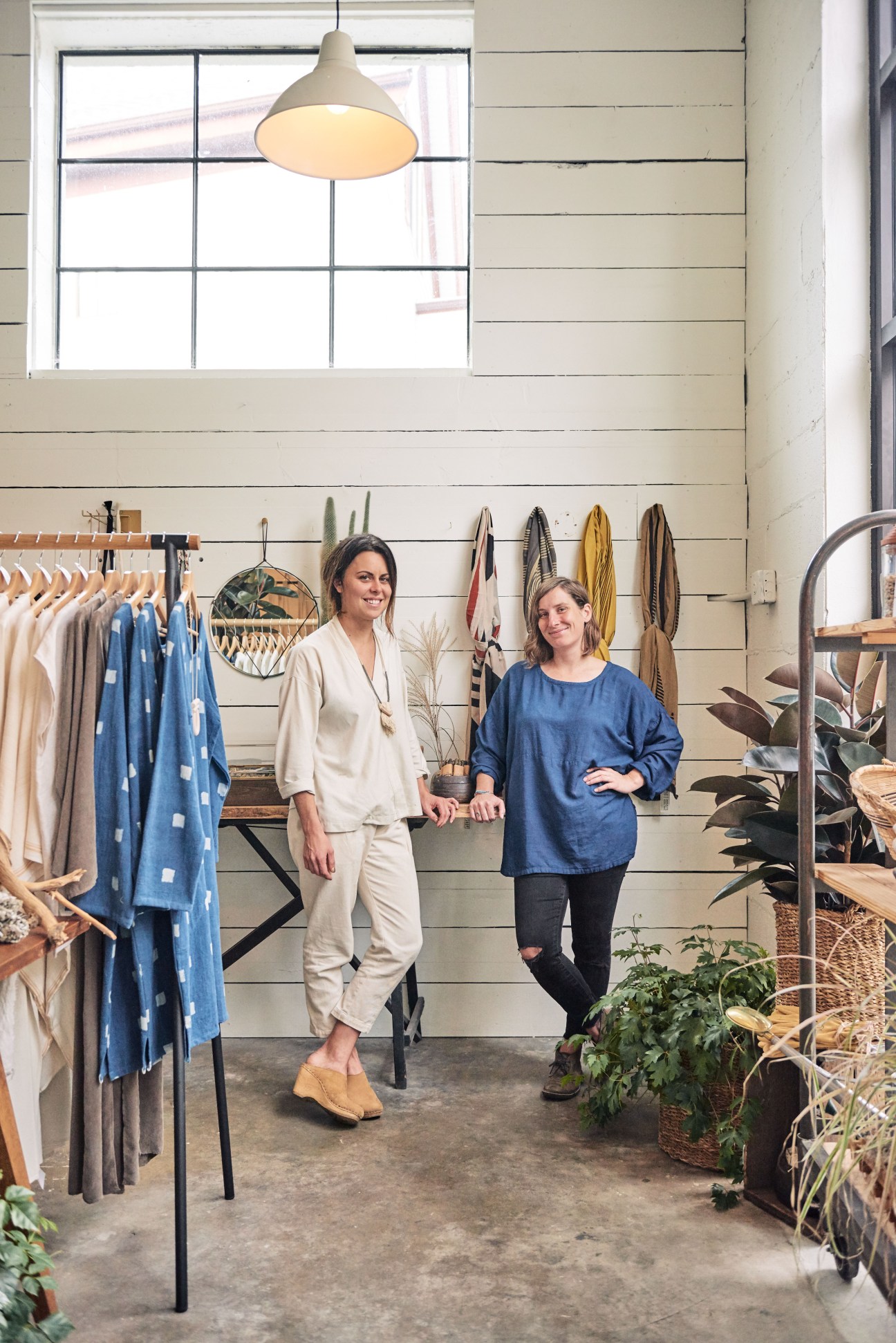 Two women stand in a retail shop with white shiplap walls, clothing racks, and hanging textiles displayed behind them.
