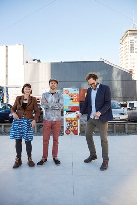 Three people stand on a temporary dance floor in an urban setting with industrial buildings in the background.