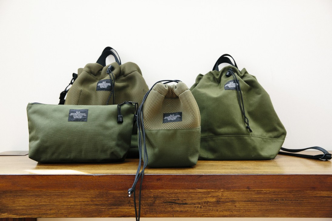 Three olive green canvas tote bags with black handles arranged on a wooden shelf.