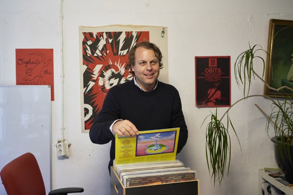 Man in dark sweater holding vinyl records and album artwork in a studio setting.
