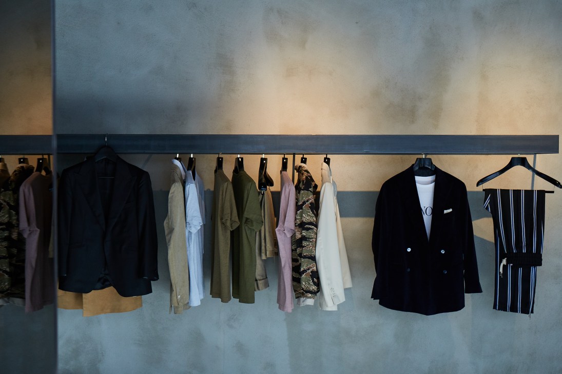 Clothing displayed on a black rail rod against a textured wall, including blazers, dresses, and patterned garments in neutral and muted tones.