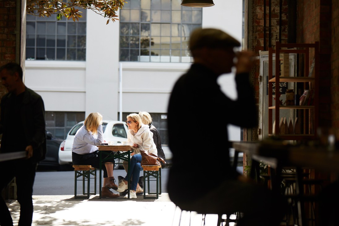 Two women seated at a table in a sunlit street-level cafe with large windows and parked cars visible outside.