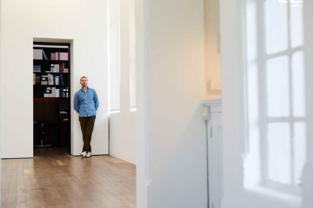 Man in blue shirt standing in minimalist interior space with black bookshelf and large windows.