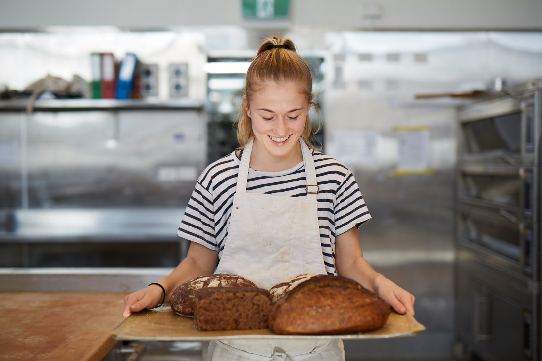Young woman wearing striped shirt and apron holding fresh loaves of bread in a bakery kitchen.