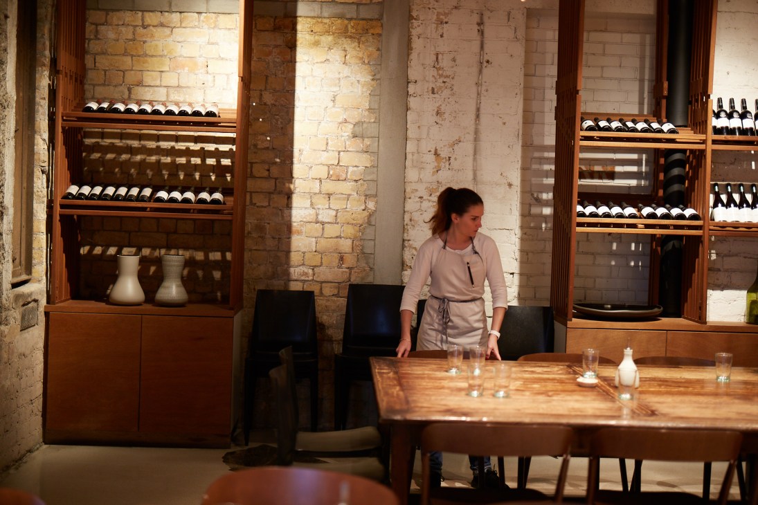 Woman standing at a wooden dining table in Amano restaurant with wine bottles displayed on wooden shelves behind her.