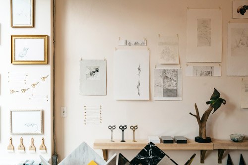 Light-filled Martha Mae shop interior displaying framed artworks, sketches, scissors, and art supplies on shelves and walls.