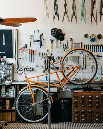 Orange vintage-inspired bicycle displayed in Heritage Bicycles General Store with tools and supplies on white pegboard wall.