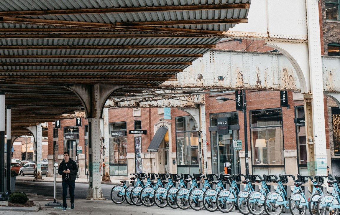 Row of bright blue Divvy bike-share bicycles lined up under a metal overpass with brick storefronts behind.