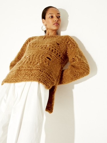 Woman wearing a tan knitted poncho over a white dress with hoop earrings.