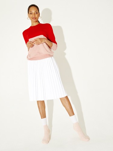 Woman wearing red cropped sweater, pink waist bag, white pleated skirt, and pale pink socks.