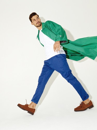 Man wearing green jacket, white shirt, blue trousers and brown suede boots jumping dynamically.