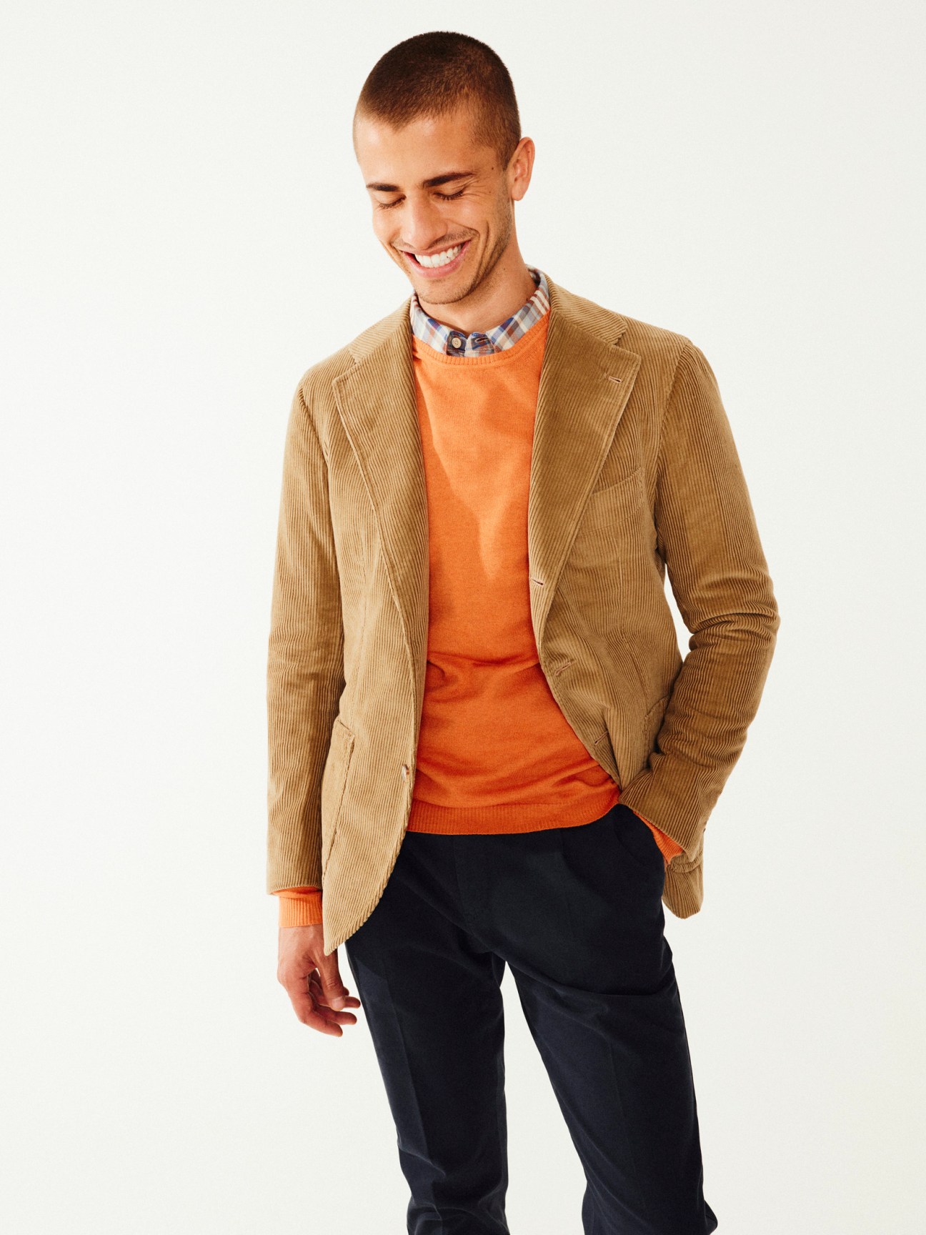 Young man wearing a tan corduroy blazer, orange crewneck sweater, and checked shirt layered for autumn styling.