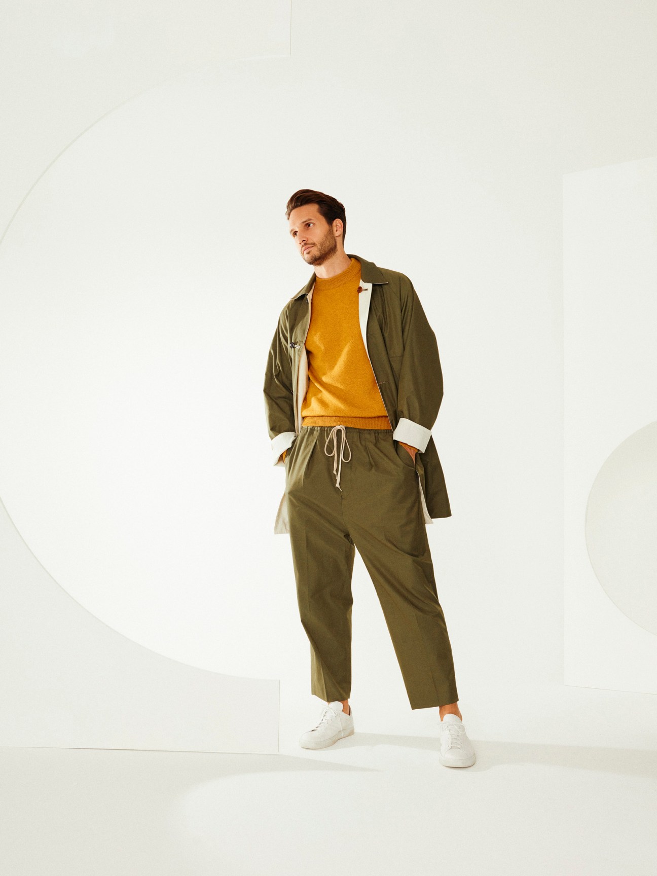 Man wearing olive-green coat and mustard-yellow sweater with white sneakers against neutral background.