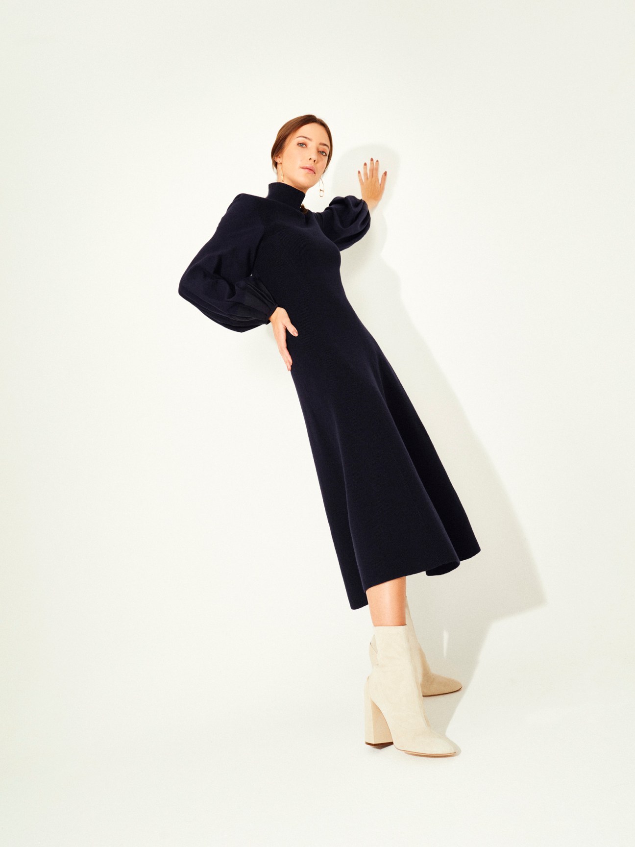 Woman wearing navy turtleneck dress with puffed sleeves and cream boots against white background.