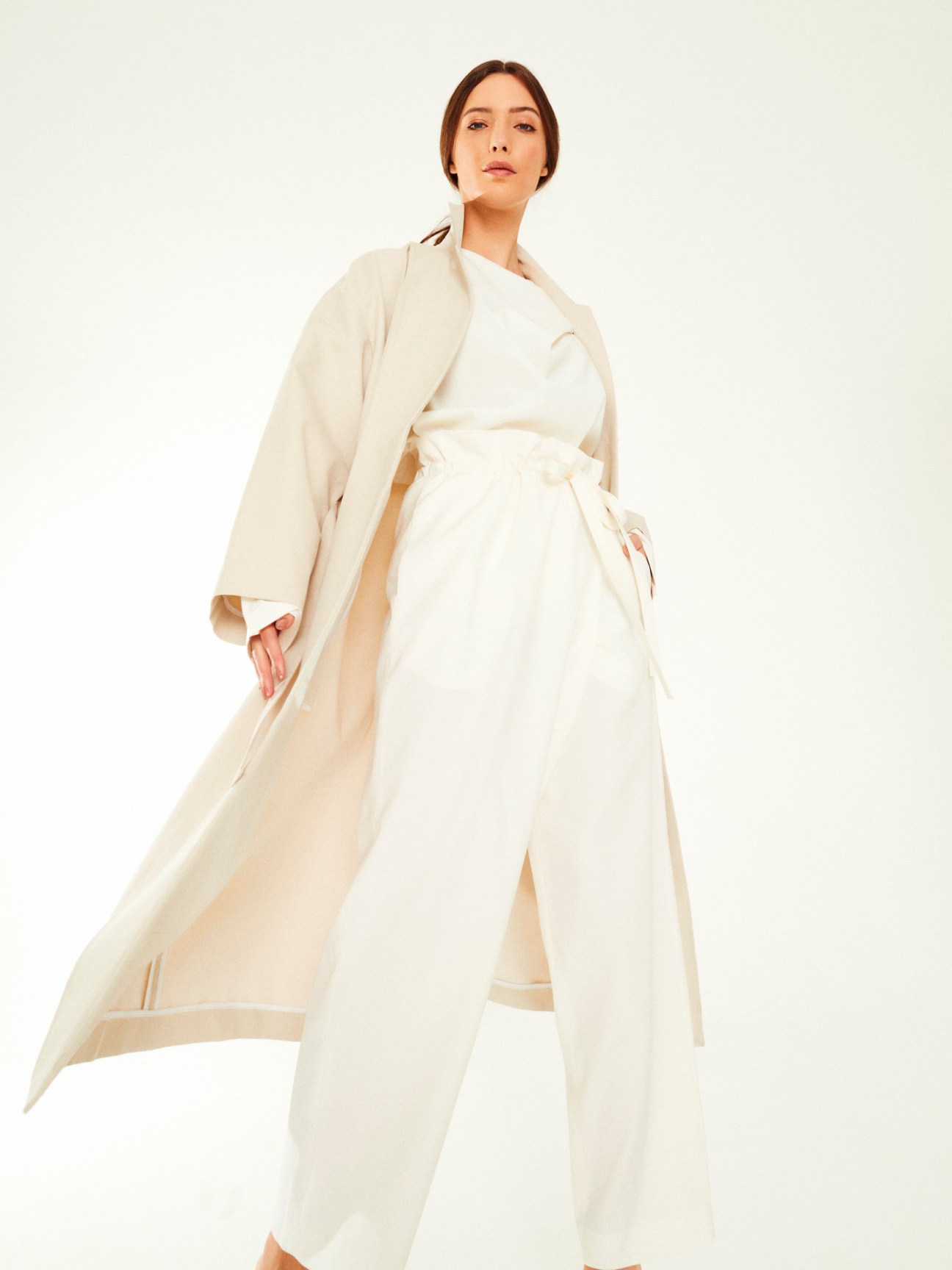 Woman wearing cream wrap coat and white wrap dress against neutral background.
