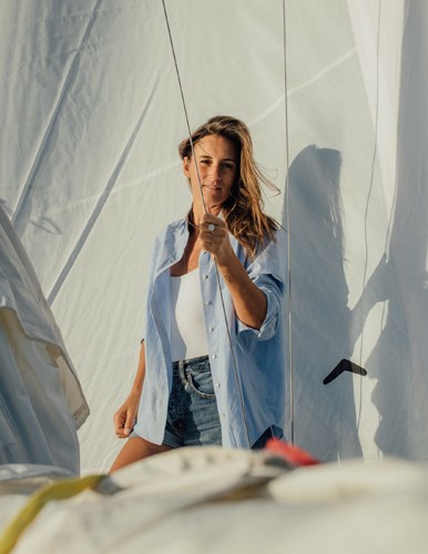 Woman in light blue shirt and denim shorts poses on a sailboat with rope and canvas visible behind her.