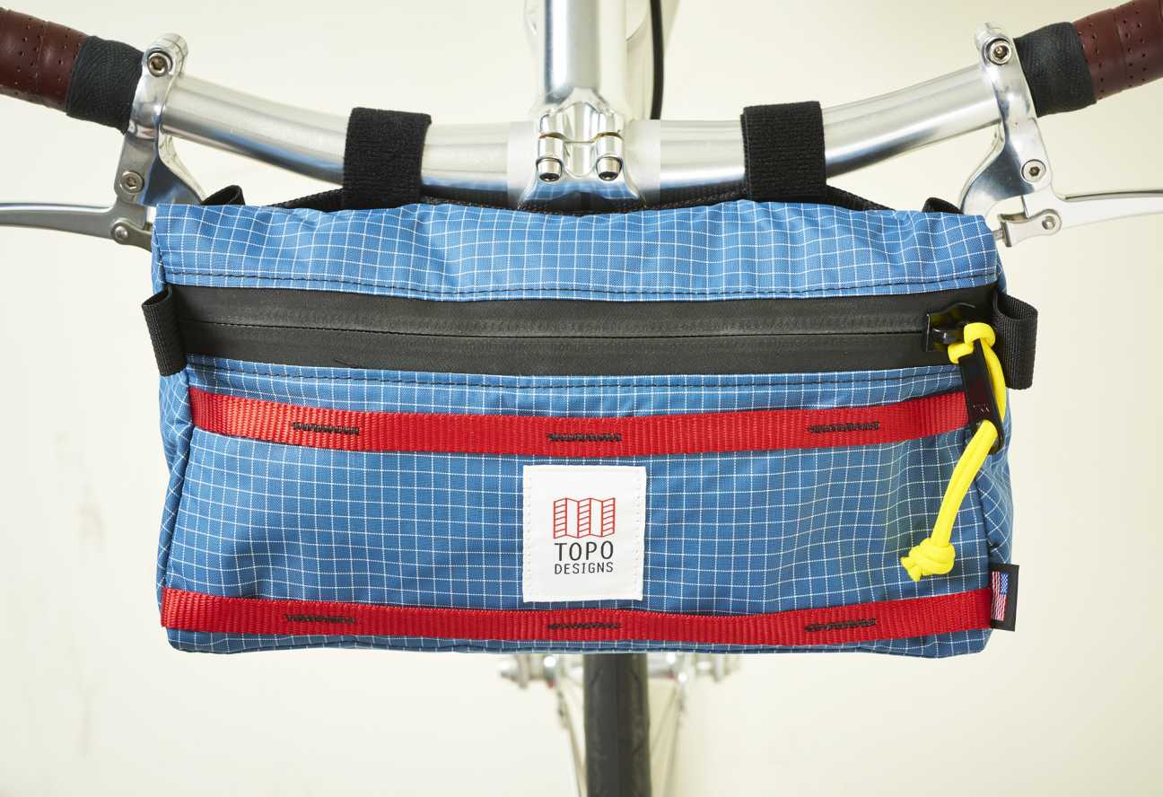 Topo Designs blue handlebar bag with red straps and yellow carabiner attached to bicycle.