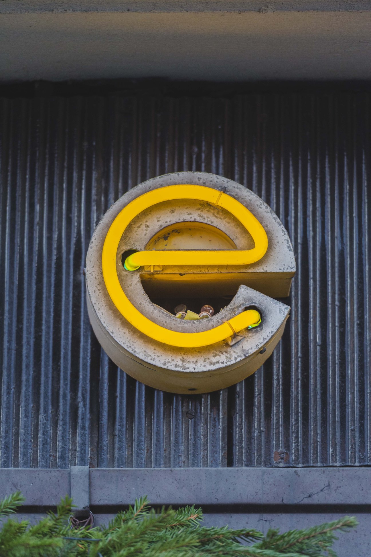 Yellow neon letter 'e' glowing against a weathered concrete and corrugated metal wall.