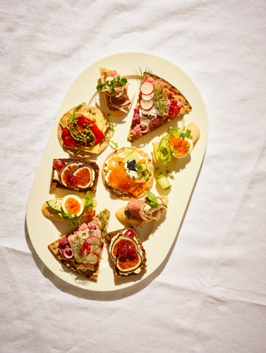 Swedish-style canapés arranged on a white plate featuring various toppings including smoked salmon, figs, roast beef, asparagus, and cheese.