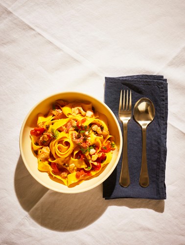 Bowl of red mullet pasta with golden ribbon noodles, red sauce, and crispy capers, served with gold cutlery on a dark napkin.