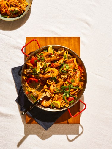 Paella with seafood, chicken, and vegetables in a black pan with red handles on a wooden board.