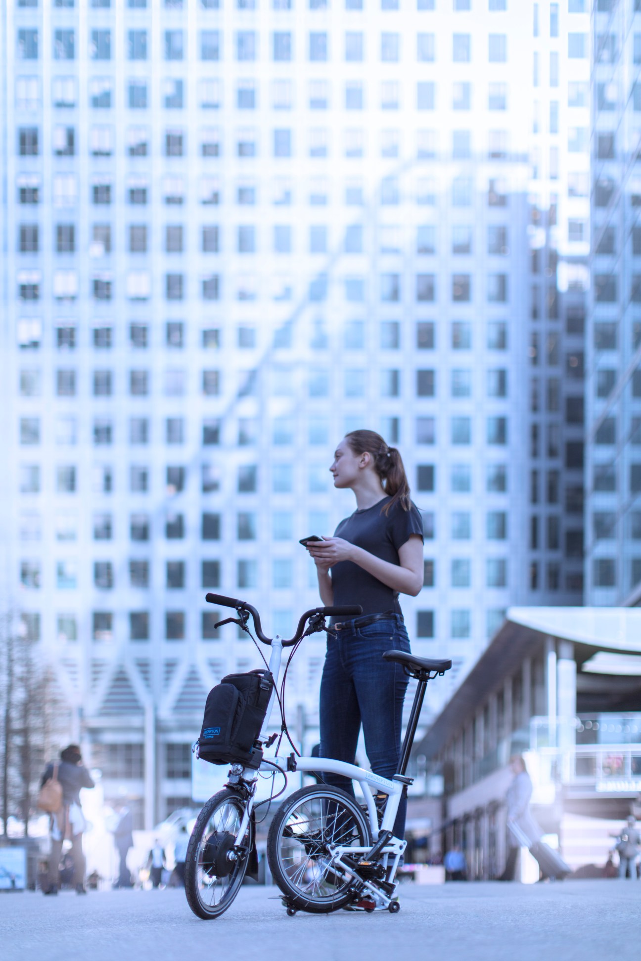 Woman standing on white electric bike with backpack in urban plaza with tall buildings behind