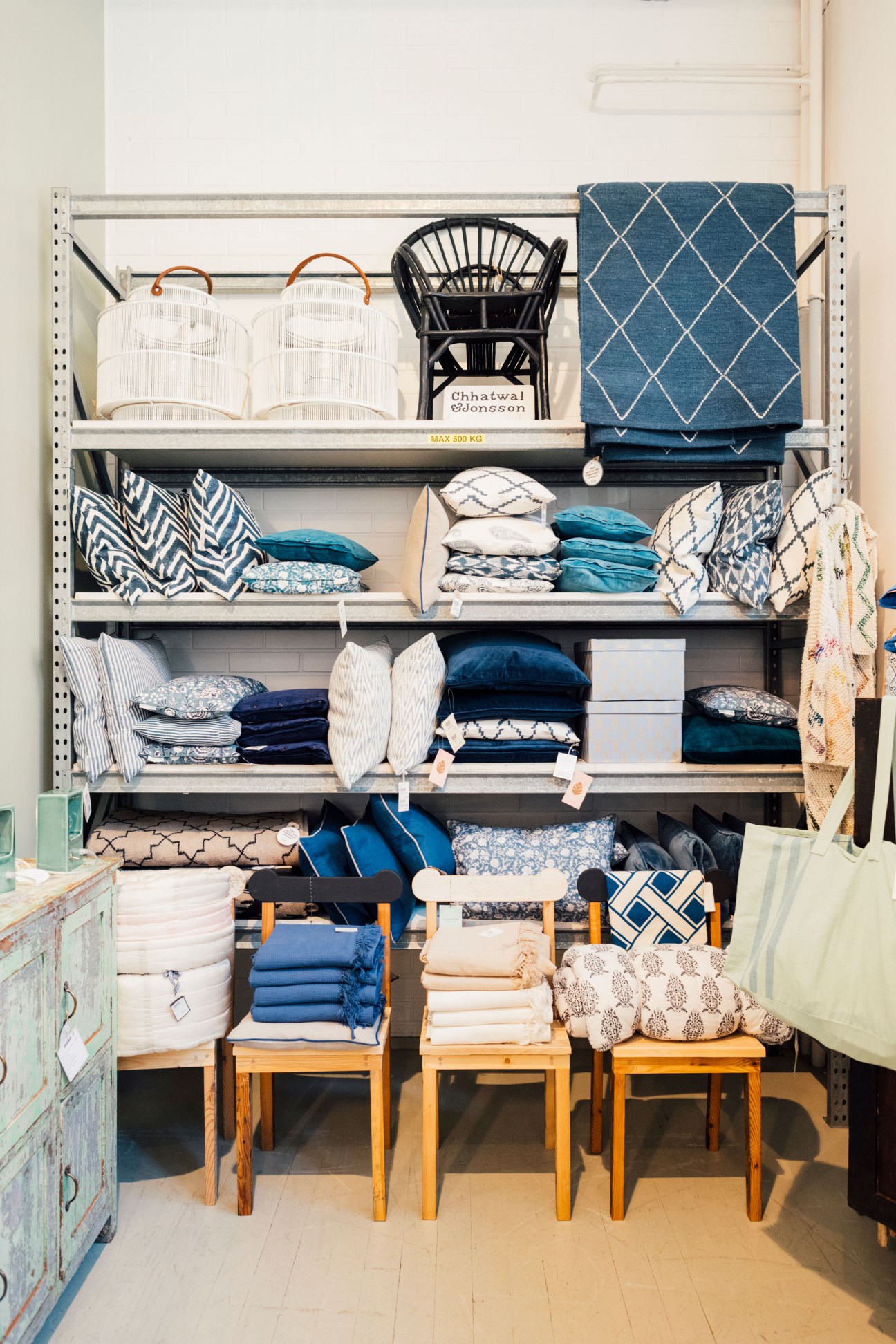 Metal shelving unit displaying folded textiles, pillows, blankets, and home goods in blue, white, and neutral tones with wooden chairs below.