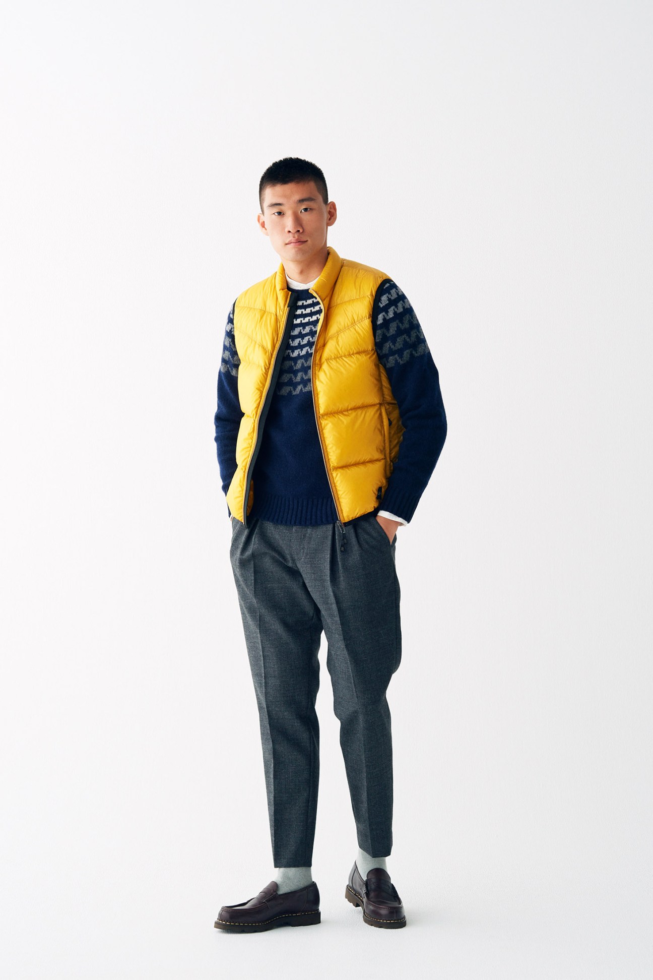 Man wearing a bright yellow quilted down vest over a dark navy knit sweater, paired with grey trousers and dark shoes.