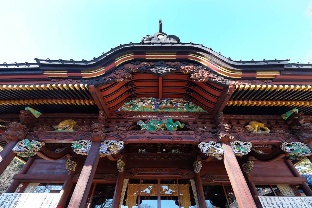 Traditional Japanese temple facade with ornate curved roof, red pillars and colorful decorative carvings against blue sky.