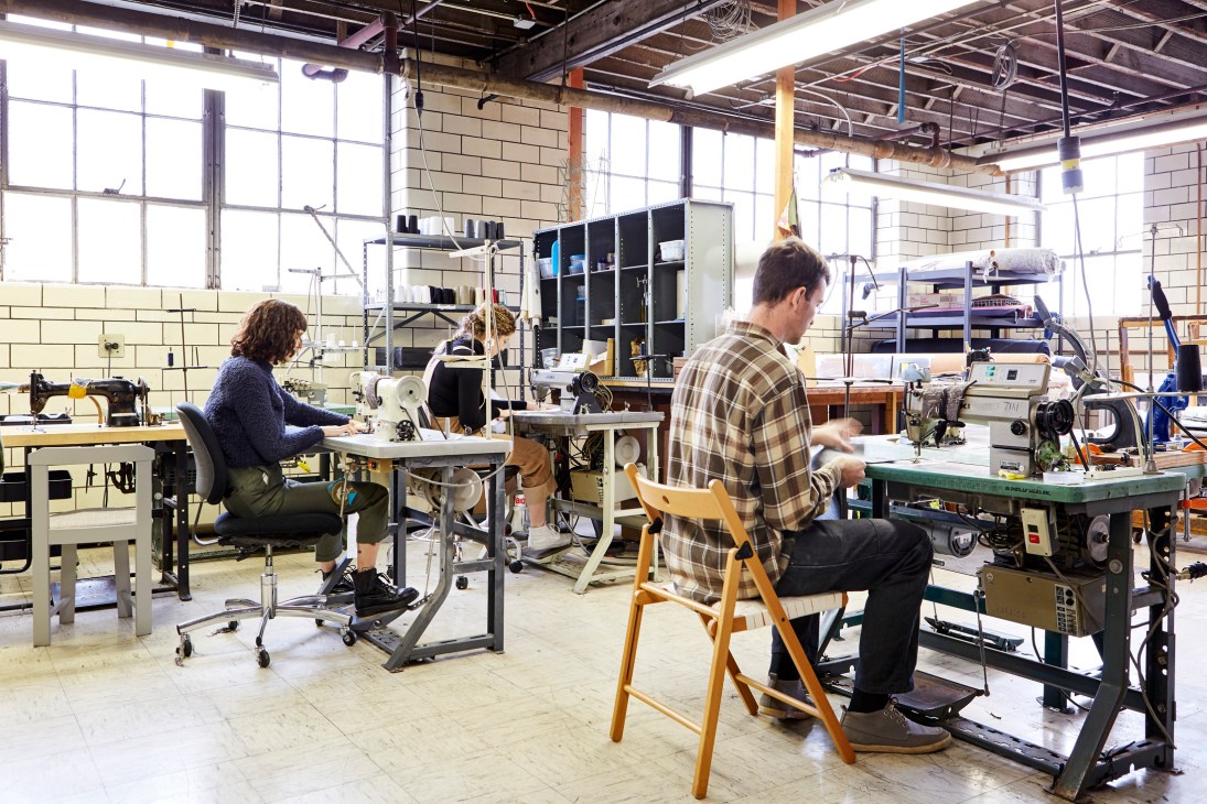 Two designers work at sewing machines in a bright industrial studio space with large windows and exposed brick walls.