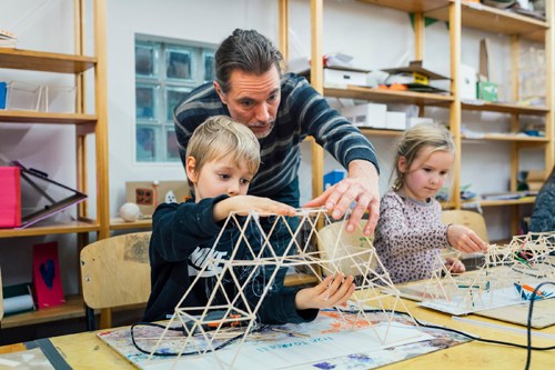 An adult and two children building a wooden structure together at a table with craft materials.