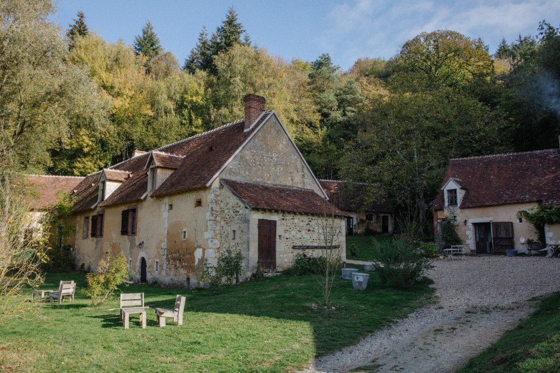 Rustic 17th-century farm buildings with stone and cream-colored walls surrounded by autumn foliage and forest.