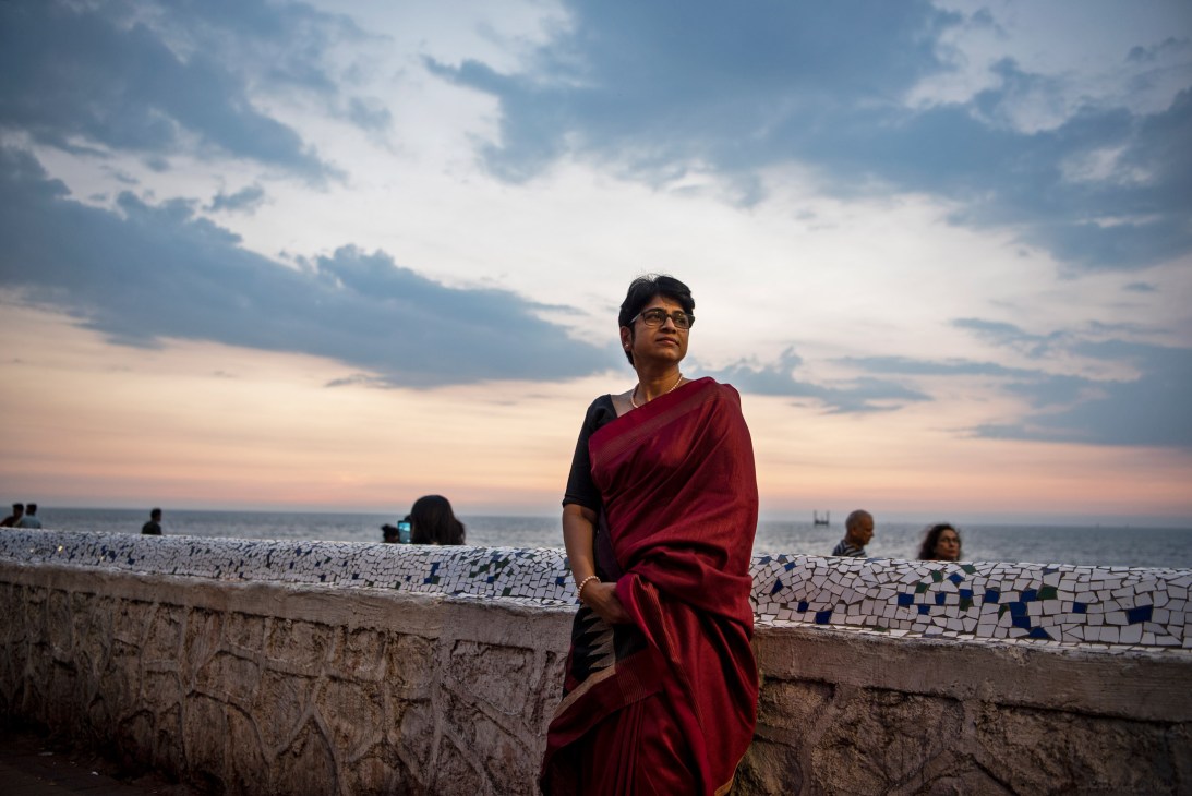 Woman in red sari standing by coastal wall at sunset with ocean and beach in background.