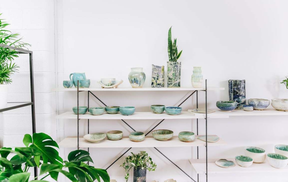 Shelving unit displaying ceramic pottery bowls, planters, and vases in green and blue glazes with potted plants.