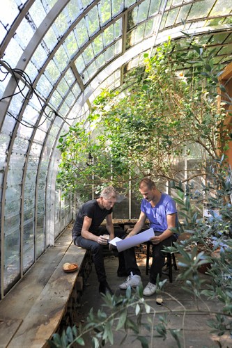 Two people work at a desk in a lush botanical garden workspace with plants climbing overhead structures.