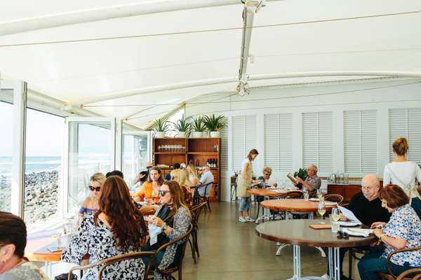 Diners seated at tables in a bright restaurant with floor-to-ceiling windows overlooking water
