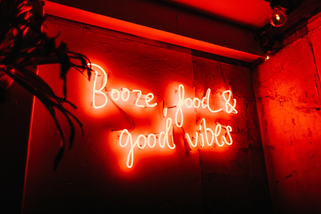 Red neon sign reading 'Booze, food & good vibes' on a brick wall