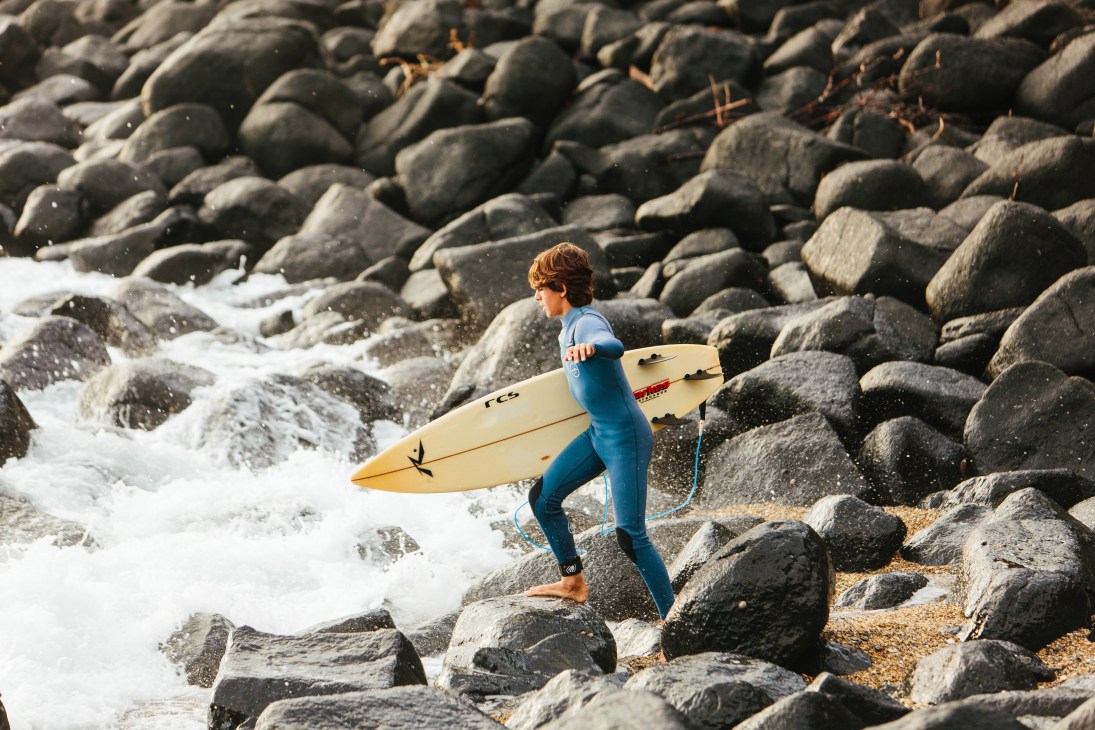 A surfer in a blue wetsuit carrying a surfboard walks across rocky coastline with crashing waves