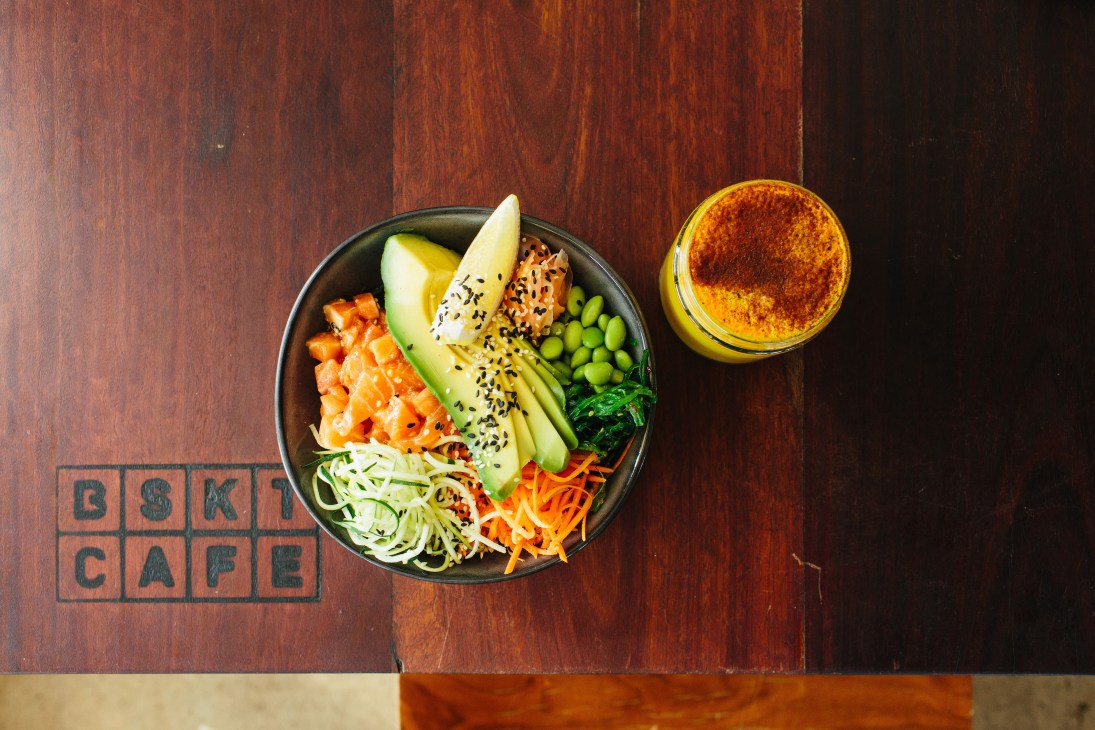 A colorful poke bowl with salmon, avocado, edamame and vegetables next to a turmeric latte at BSKT cafe