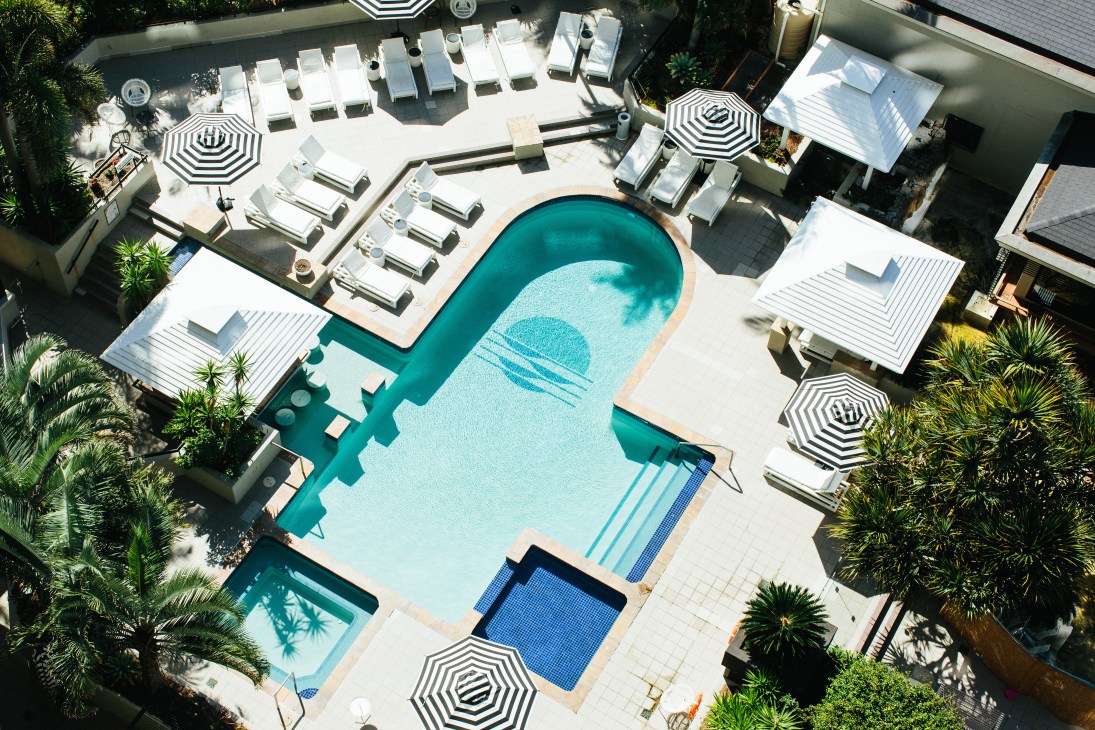 Aerial view of QT Gold Coast hotel pool area with white loungers, striped umbrellas and tropical landscaping