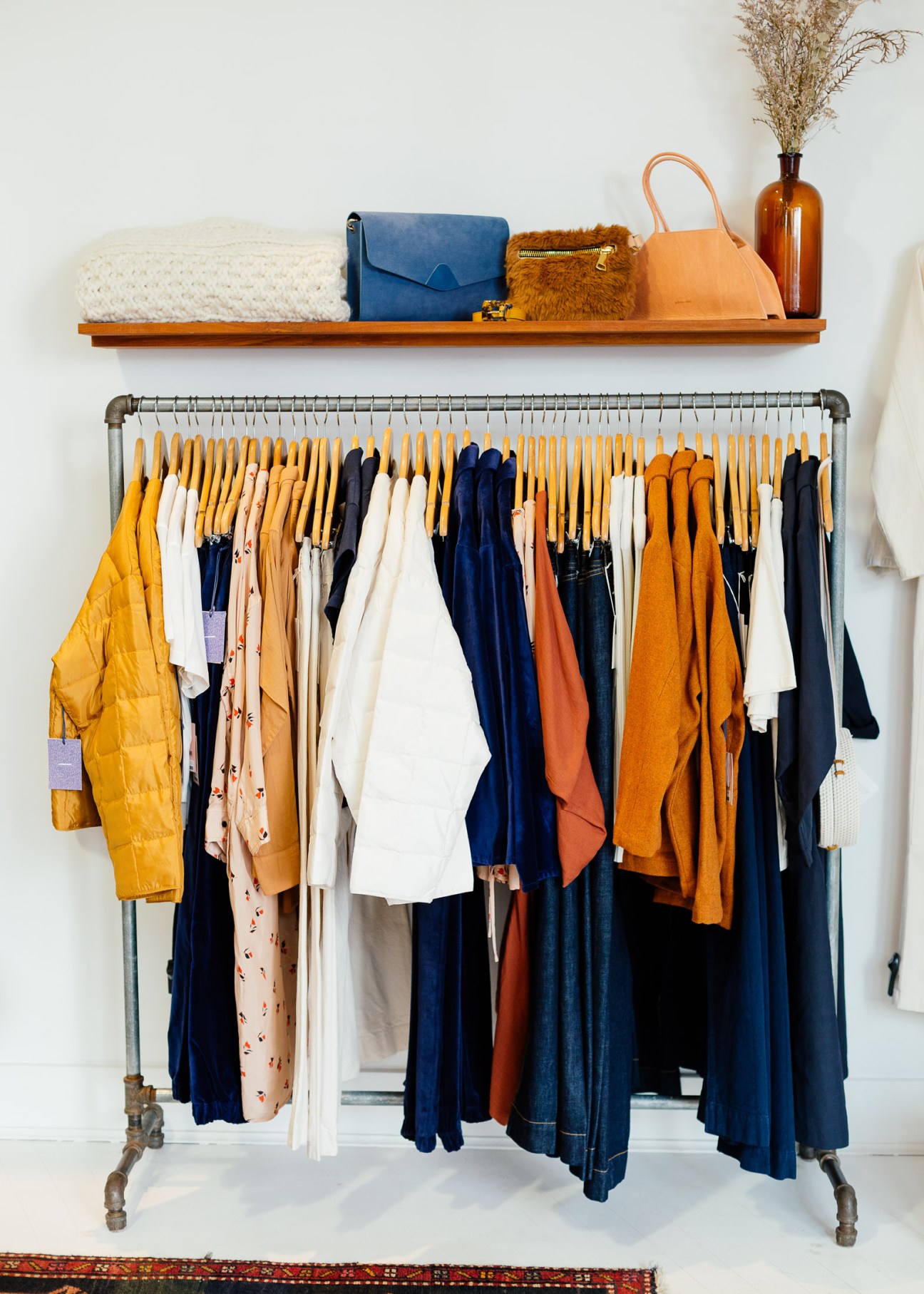 Clothing rack displaying organized wardrobe with yellow, navy, white and orange garments on hangers, with accessories and bags on wooden shelf above.