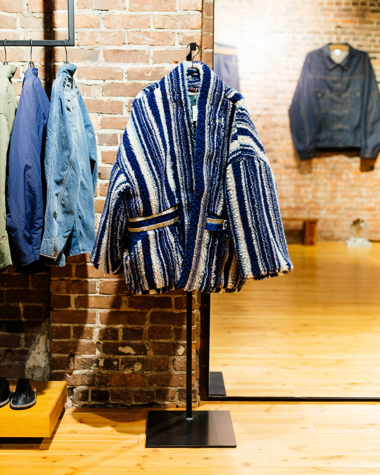 Blue and white striped wool cape hanging on a stand against an exposed brick wall with denim jackets in the background.