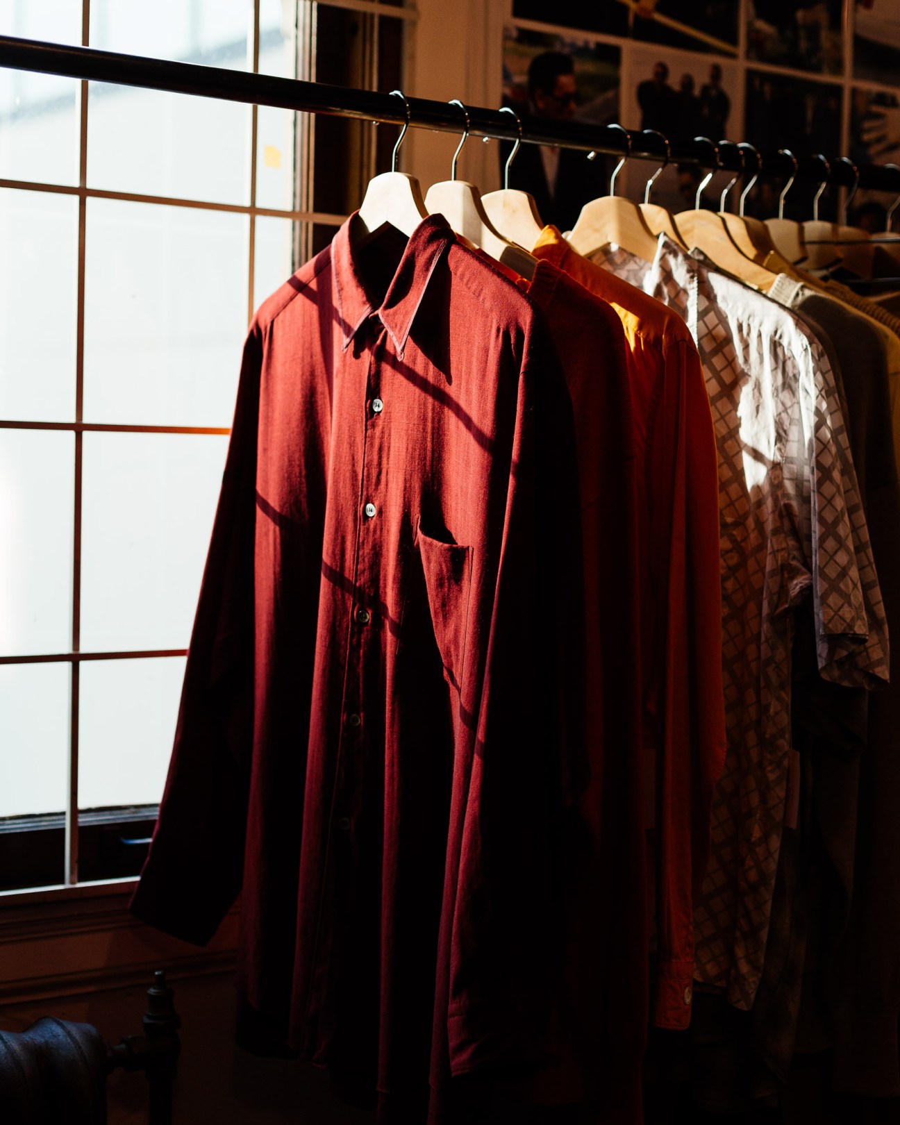 Red button-up shirt and other vintage clothing hung on a black rod against a window.