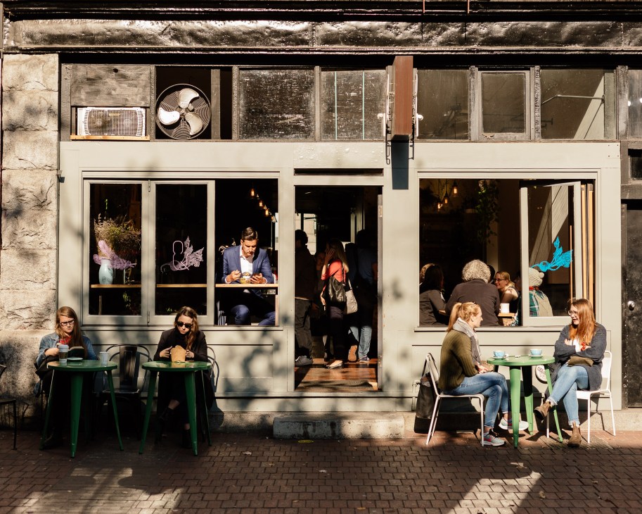 Customers seated at green tables outside a café with large windows displaying the interior.
